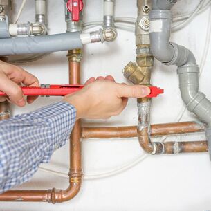 Plumber in blue plaid shirt using red wrench to adjust copper pipes along wall